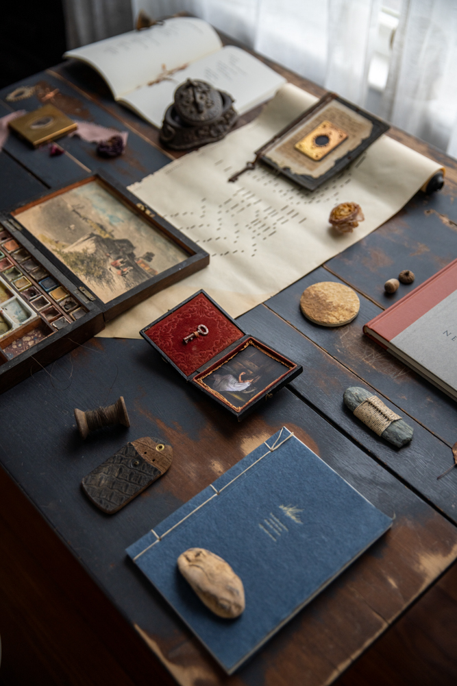 Сокровища художника и фотообъекты ручной работы, разложенные на винтажном деревянном столе, в том числе любимые книги, набор для акварели 19 века, различные найденные предметы, и природные элементы, такие как сосновые шишки и камни, найденные в штате Мэн во время прогулок. © Кристин Клафф